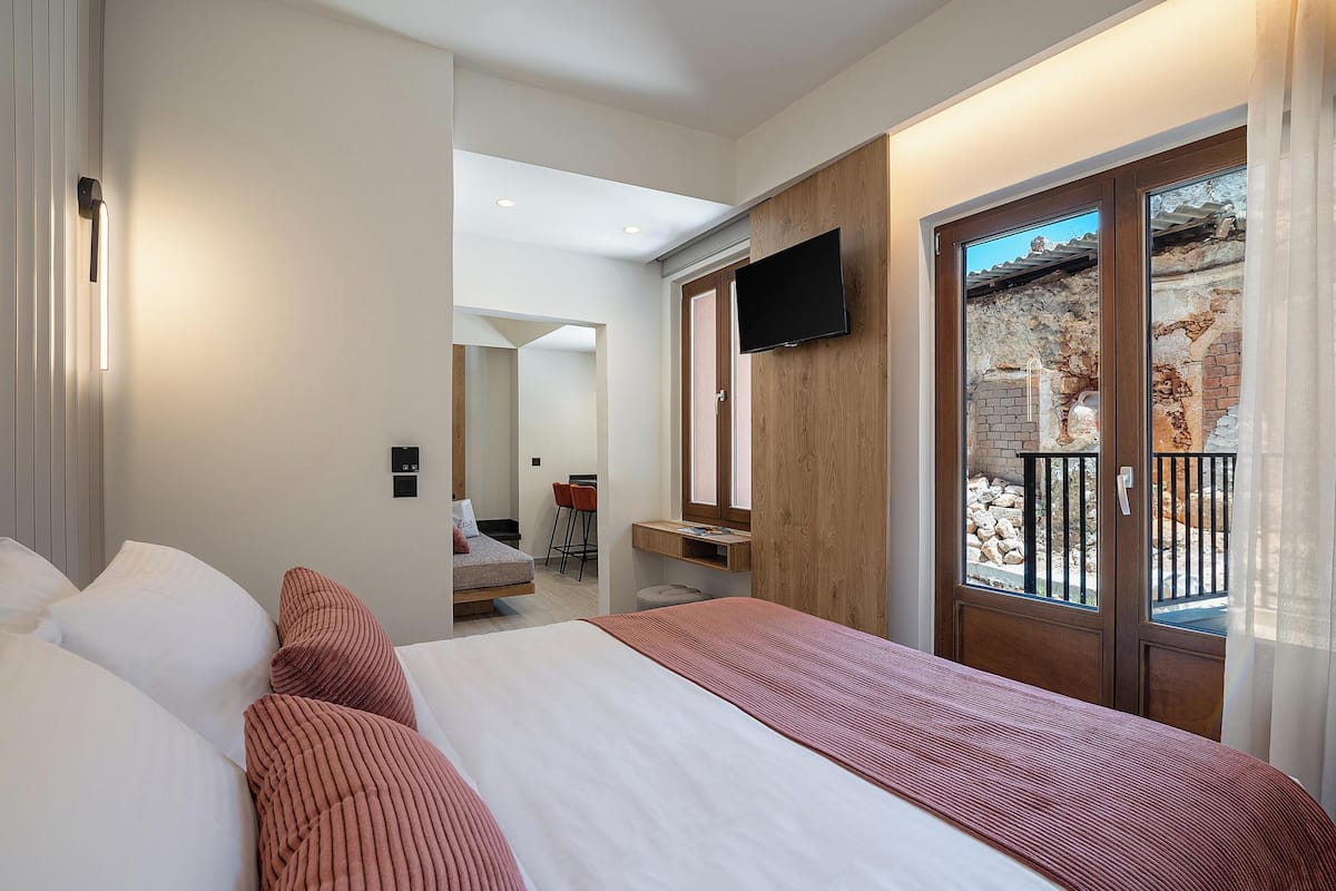 A hotel room with a bed, a TV mounted on the wall, a wooden door with a balcony, and a small desk with a chair.