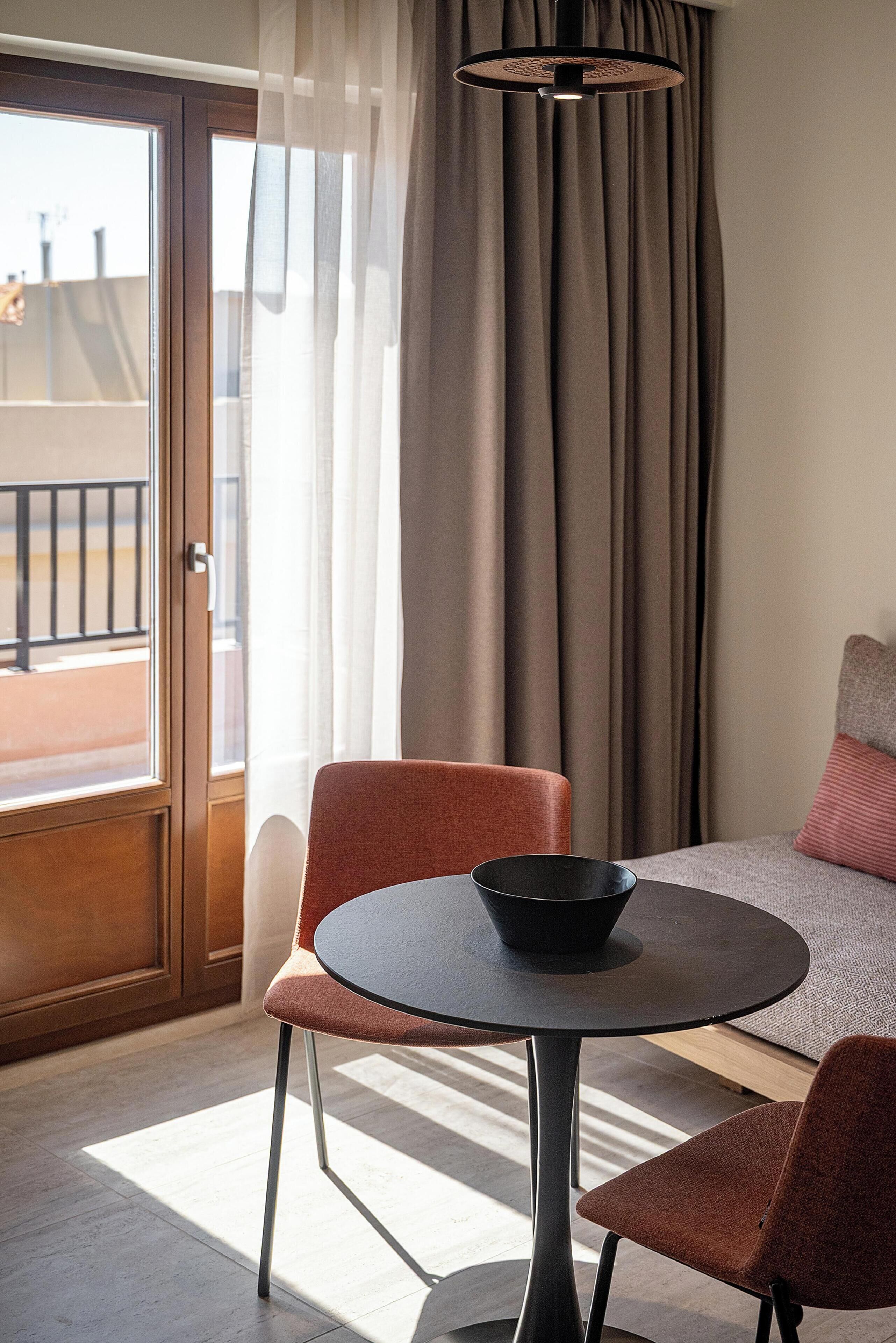 Habitación de hotel con cama, una mesa redonda con un bol, dos sillas, ventana con cortinas y vistas al balcón.