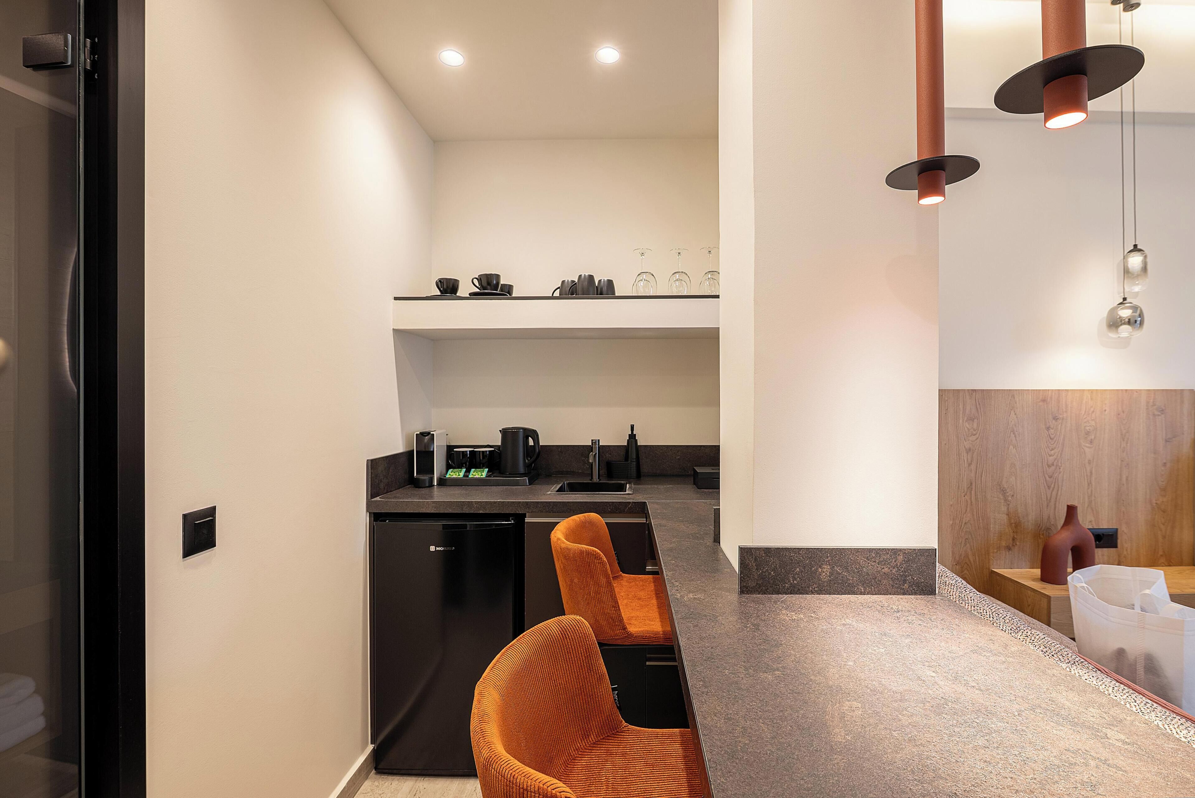 A modern kitchen with a black countertop, a coffee machine, and a kettle. There are two orange chairs in the foreground.