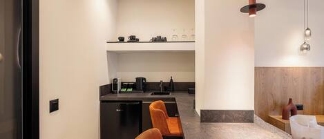 A modern kitchen with a black countertop, a coffee machine, and a kettle. There are two orange chairs in the foreground.