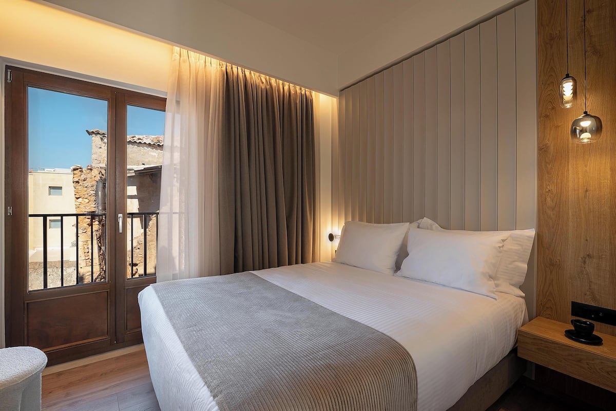 A hotel room with a large bed, a wooden nightstand, a window with curtains, and a view of buildings outside.