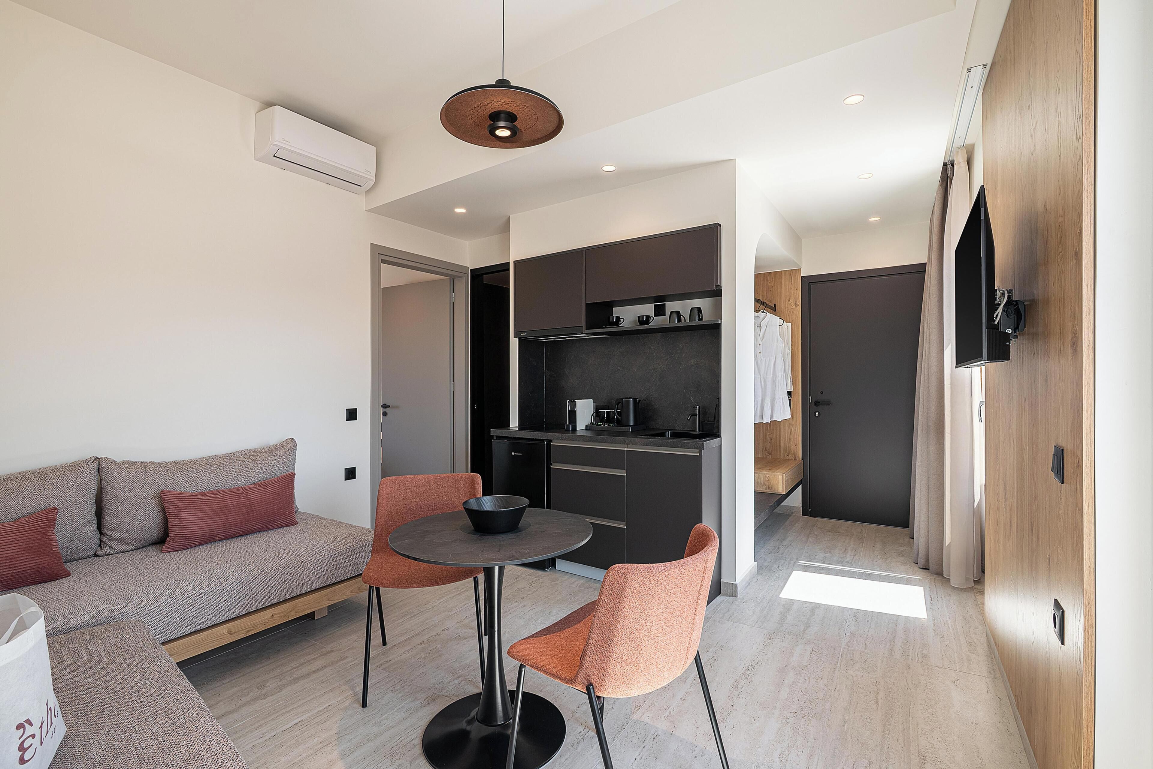 A modern living space with a sofa, two chairs, a small table, a kitchen area, and a TV mounted on the wall.