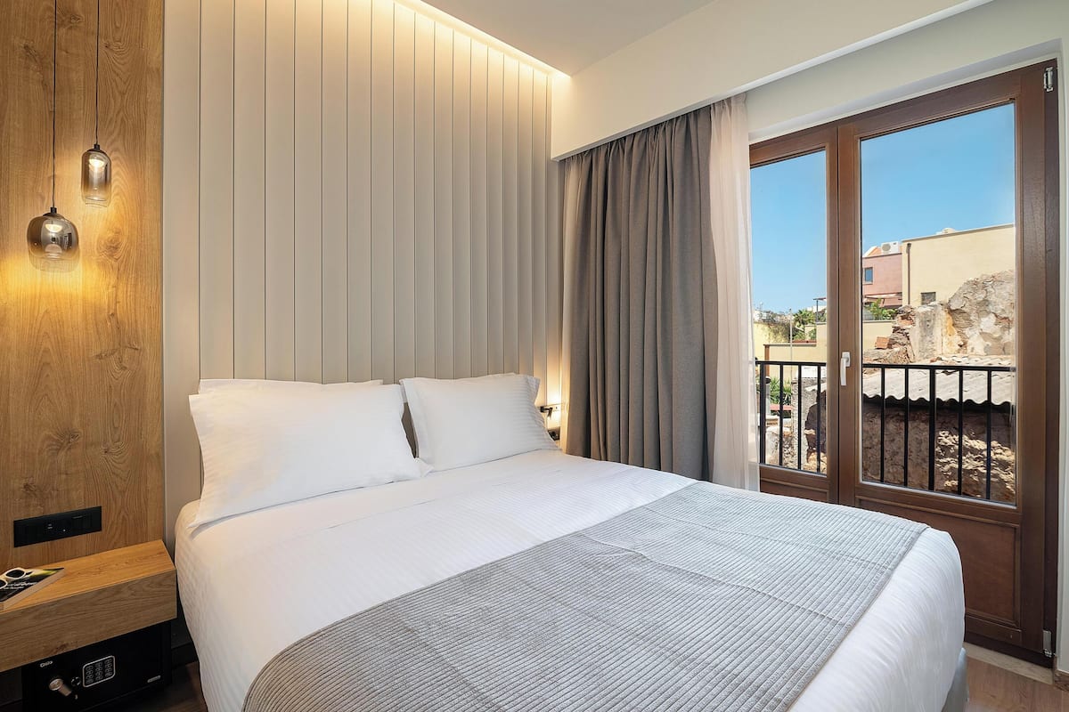 A hotel room with a large bed, a wooden panel wall, a nightstand with a control panel, and a view of the outside through a sliding door.