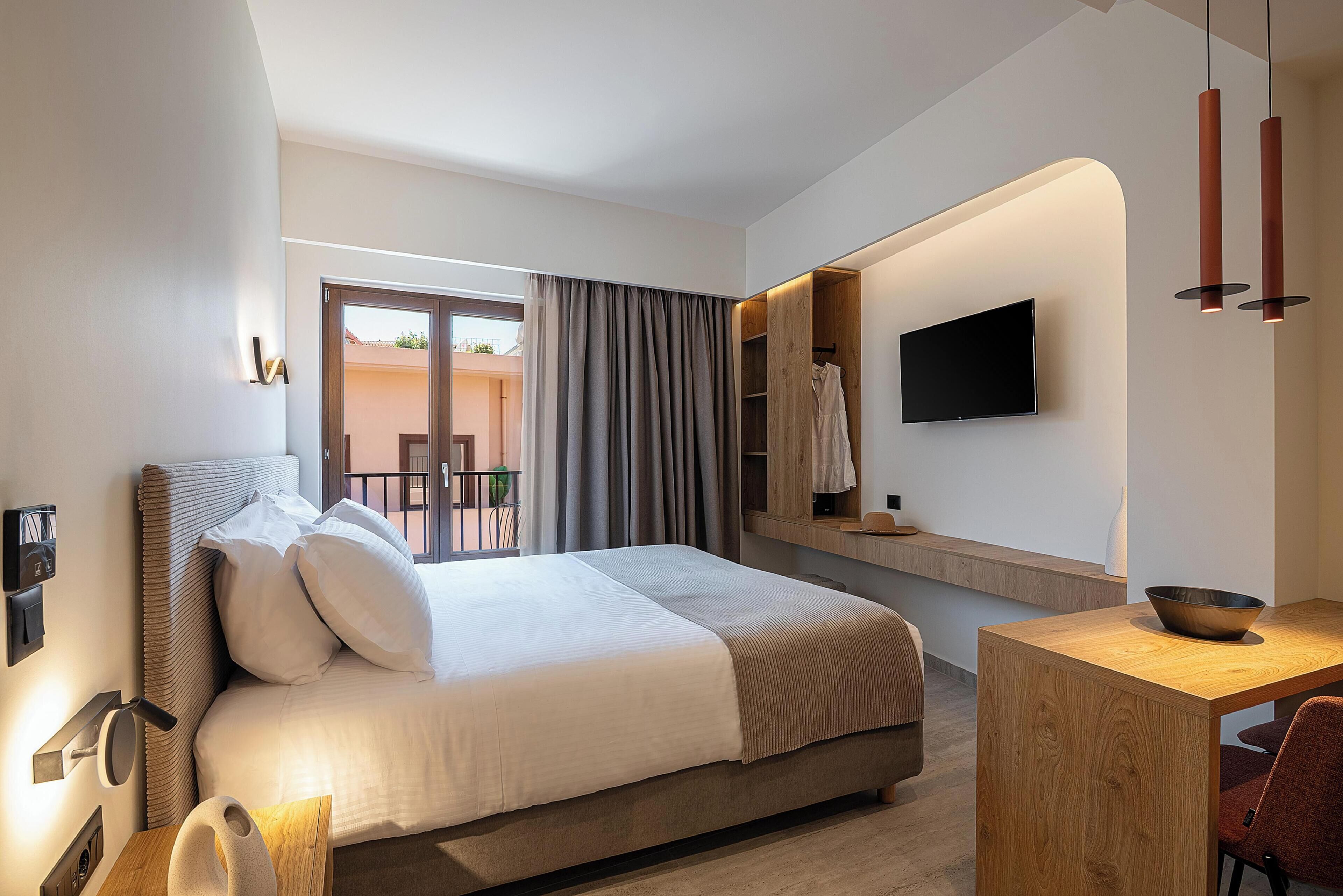 A modern hotel room with a bed, a desk, a TV, and a balcony with curtains.