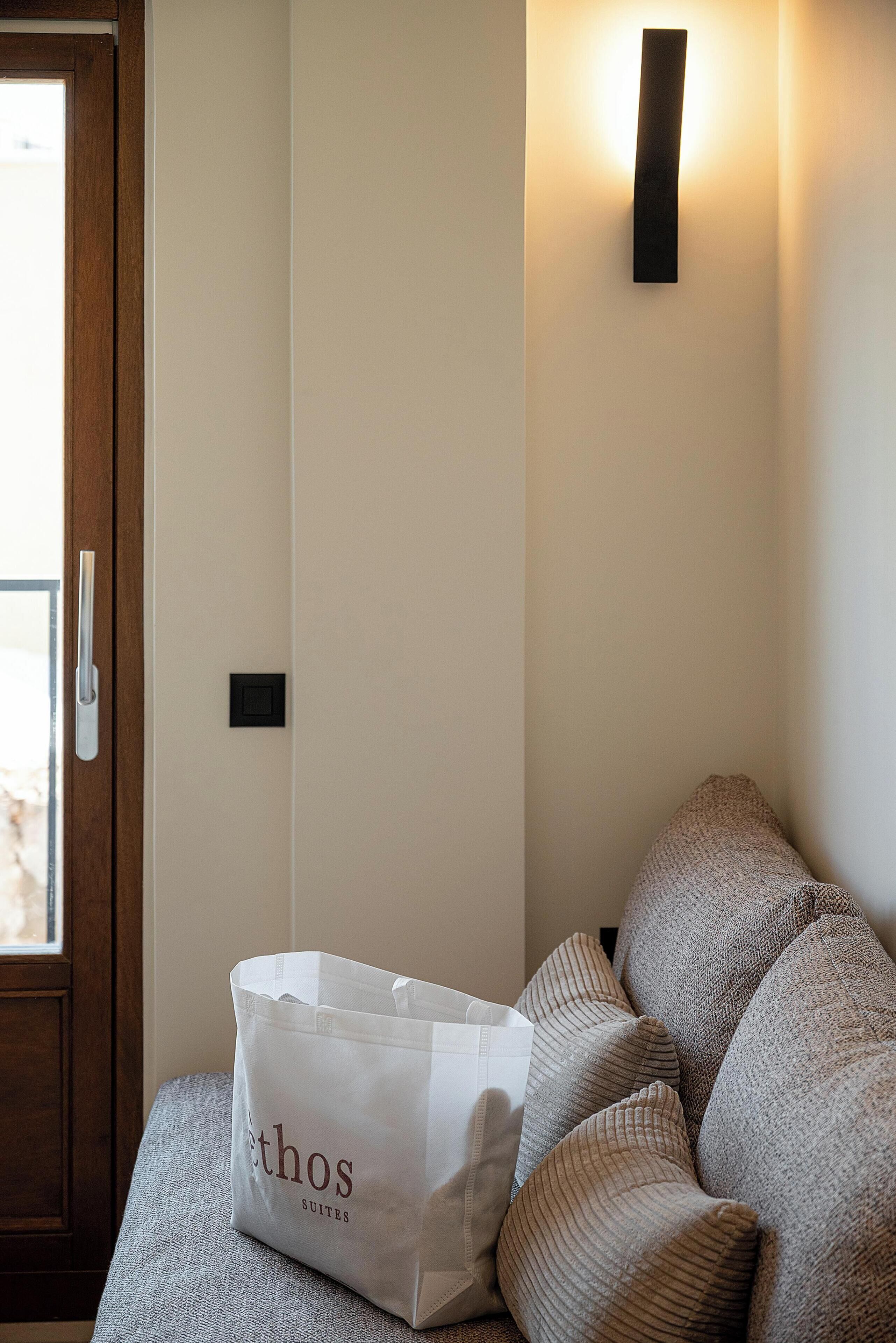 A room with a wooden door, a white bag labeled 'ethos SUITES' on a textured sofa, and a wall-mounted light fixture.