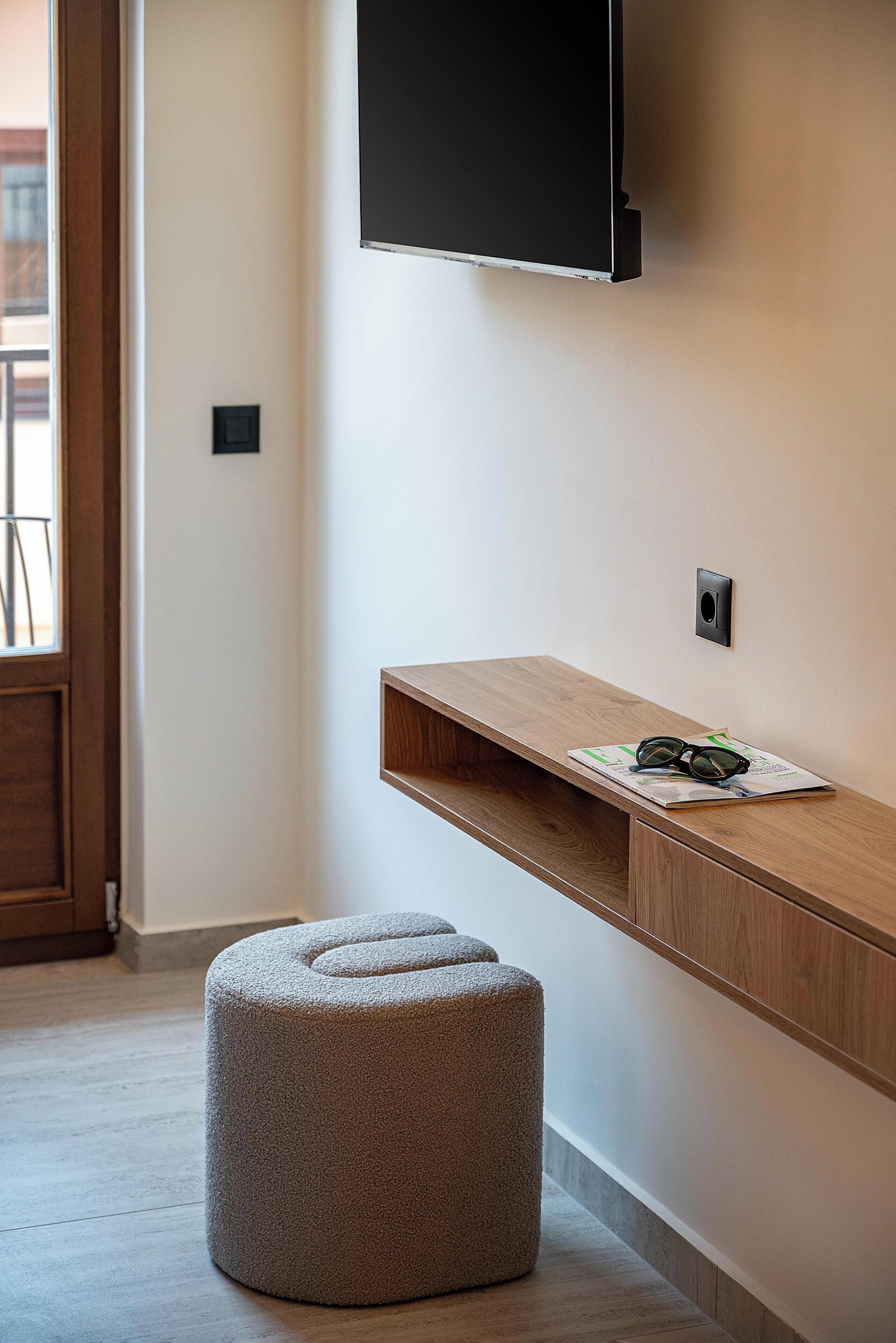 Una camera moderna con una panca in legno, un poggiapiedi rotondo, una TV a parete e una porta che dà su un altro ambiente.