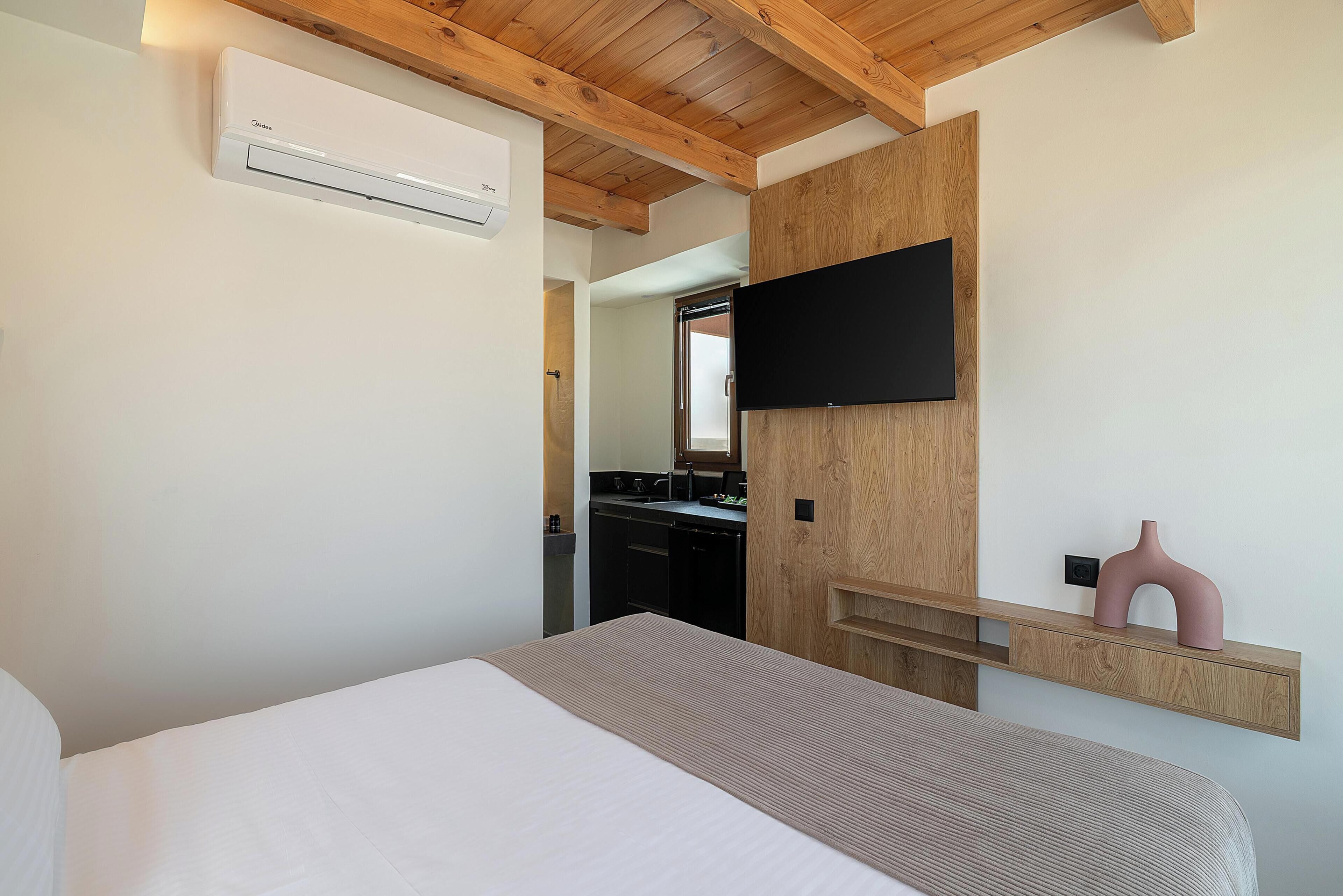 A bedroom with a bed, a mounted TV, a wooden shelf, and a kitchen area visible through an open door.