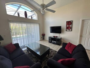 A living room with a ceiling fan, a flat-screen TV on a black stand, a glass coffee table, and dark sofas with red cushions. - The Beauty of Solana Near Disney and Universal (Davenport)