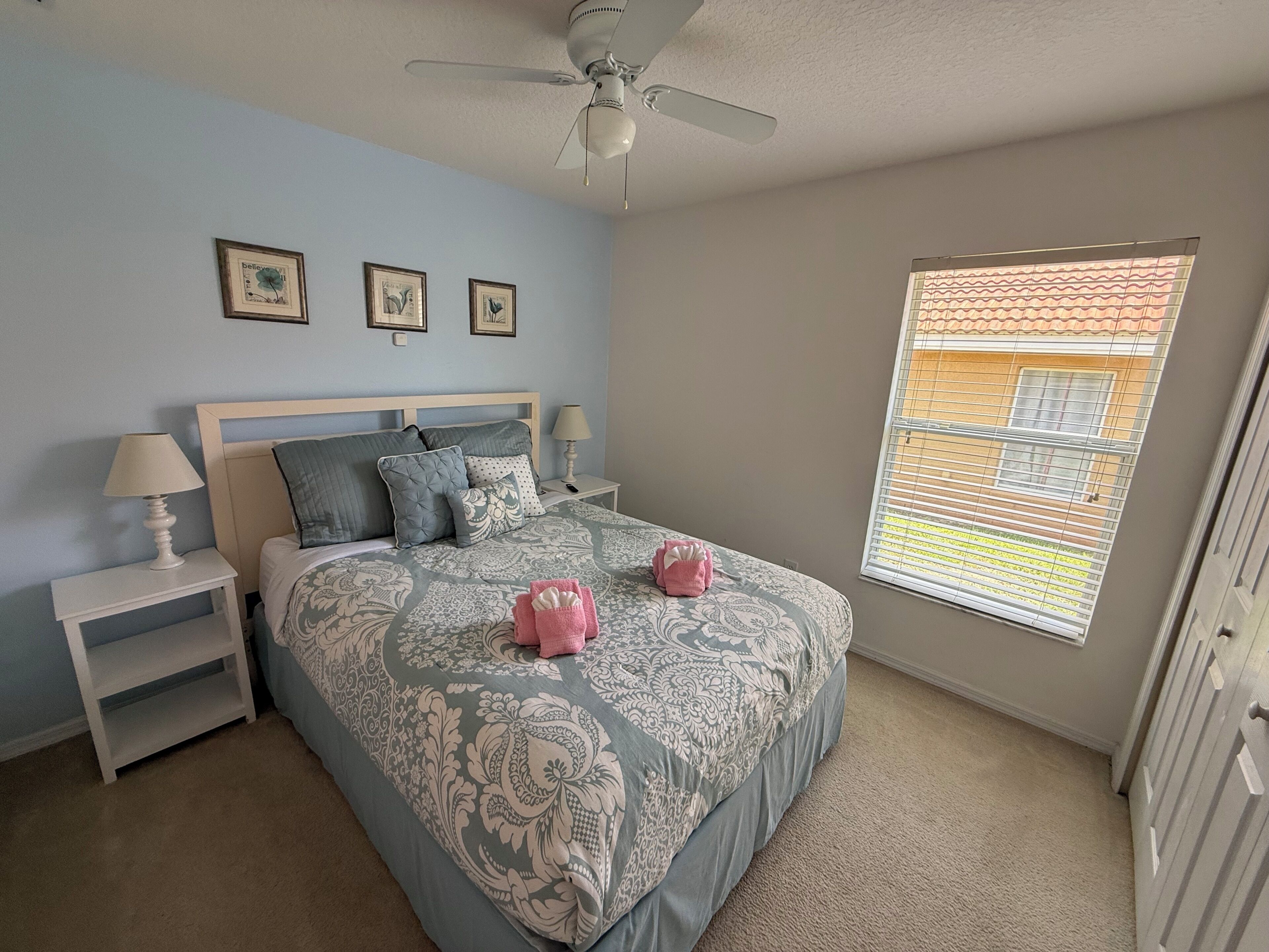 Une chambre à coucher avec un lit, des tables de chevet, un ventilateur de plafond et une fenêtre avec des stores.