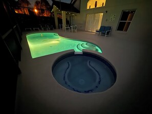 Una piscina con un jacuzzi al frente, rodeada de mobiliario de exterior y un área de patio techada.