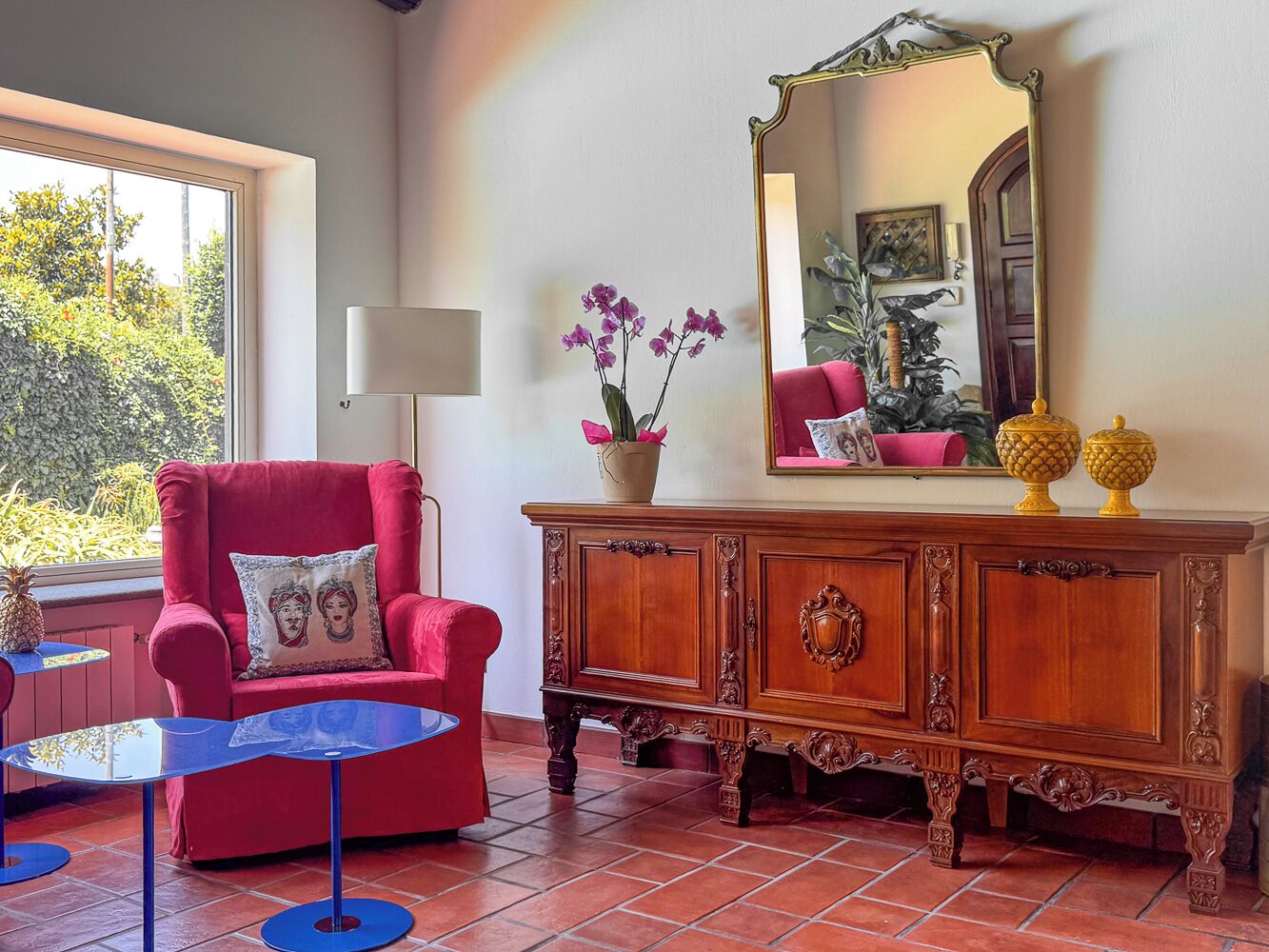 A red armchair with a cushion, a wooden cabinet with decorative elements, a mirror, and a small table with a vase of flowers.