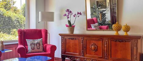 A red armchair with a cushion, a wooden cabinet with decorative elements, a mirror, and a small table with a vase of flowers.