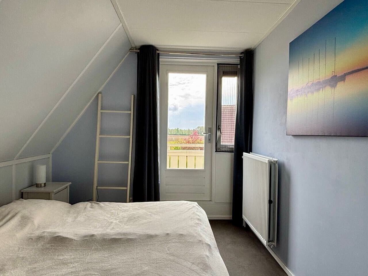 Ein Schlafzimmer mit einem Bett, einer Leiter, einem Heizkörper, einer Tür, die auf einen Balkon führt, und einem Bild an der Wand.