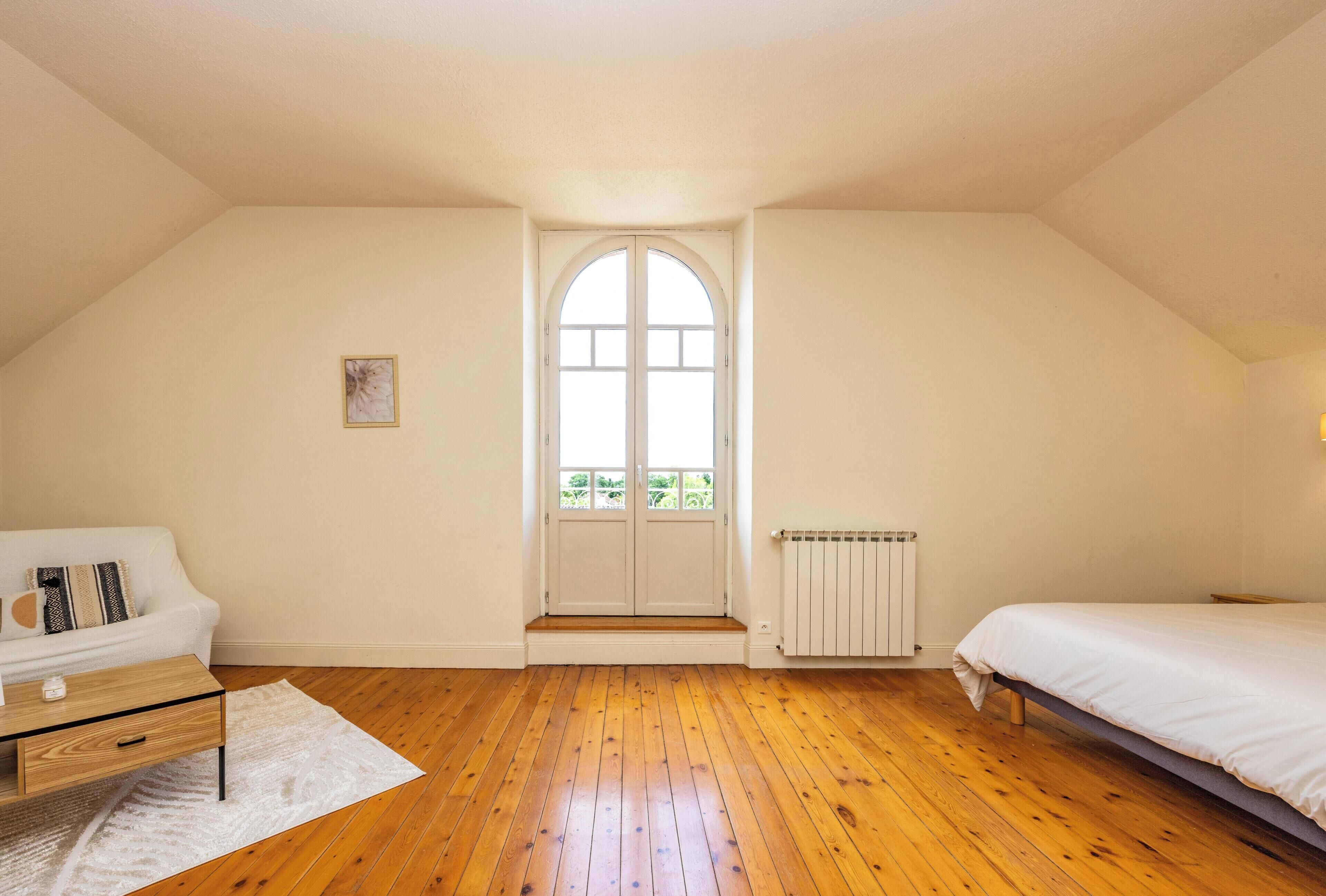 Un dormitorio con suelo de madera, una cama blanca, un sofá blanco, una mesita y una ventana con marco blanco.