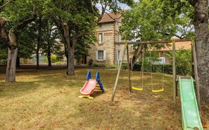 A playground with slides and swings, surrounded by trees and a stone building in the background.