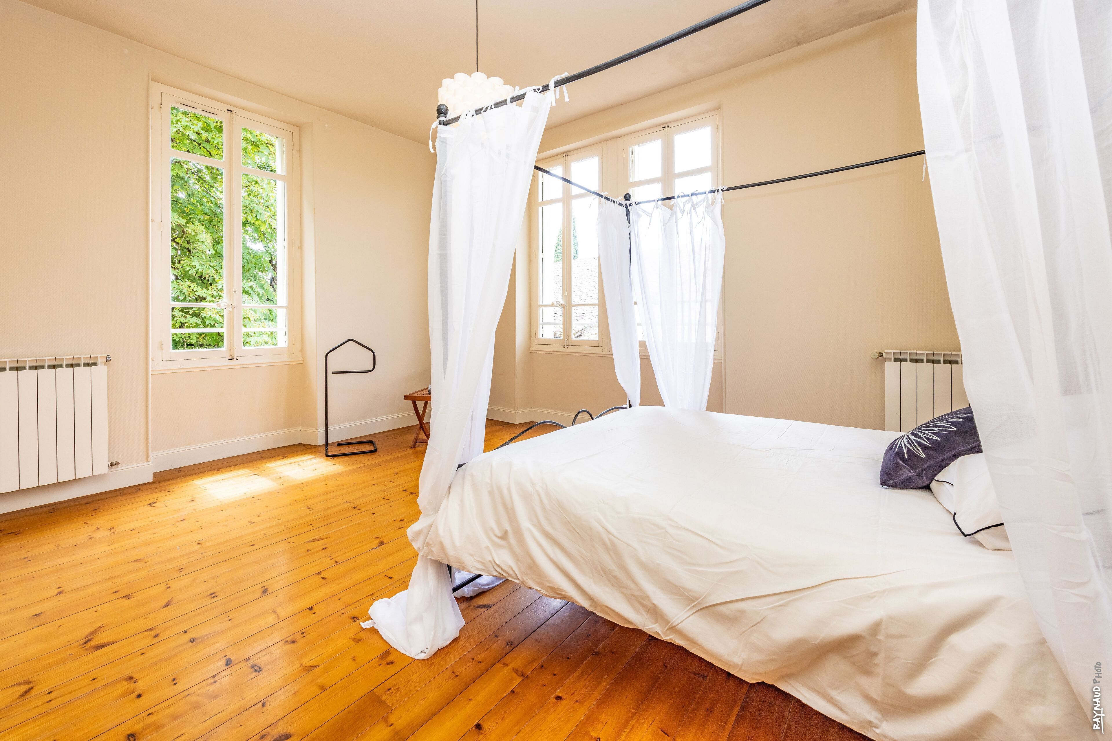 Een slaapkamer met een hemelbed, houten vloeren en uitzicht op groen buiten de ramen.