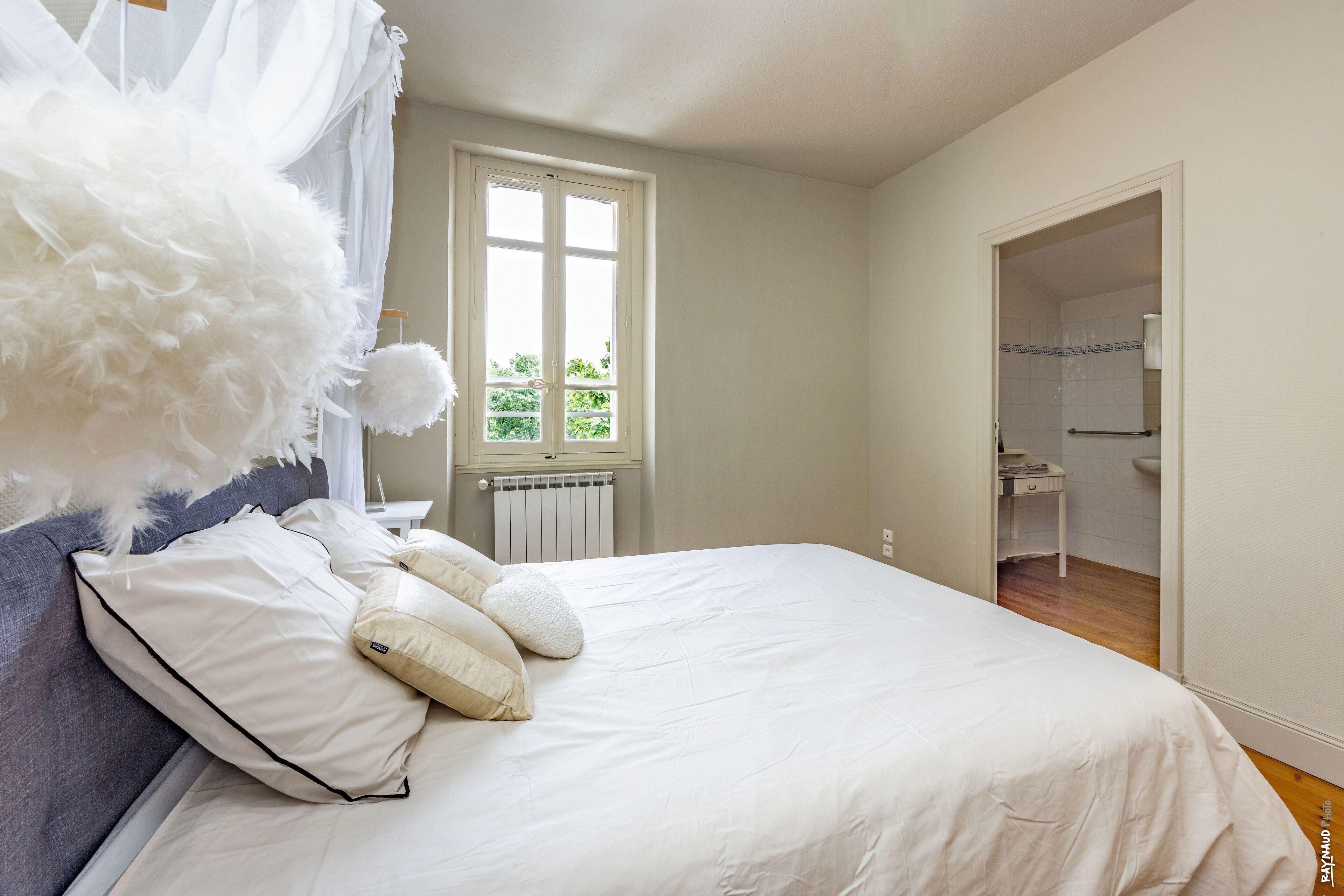 Une chambre à coucher avec un grand lit, une fenêtre avec des rideaux, un radiateur et une salle de bain visible à travers une porte ouverte.