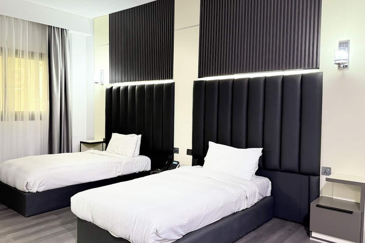 Habitación de hotel con dos camas, un ventanal grande con cortinas traslúcidas, un cabecero negro y una mesita de noche con un teléfono.