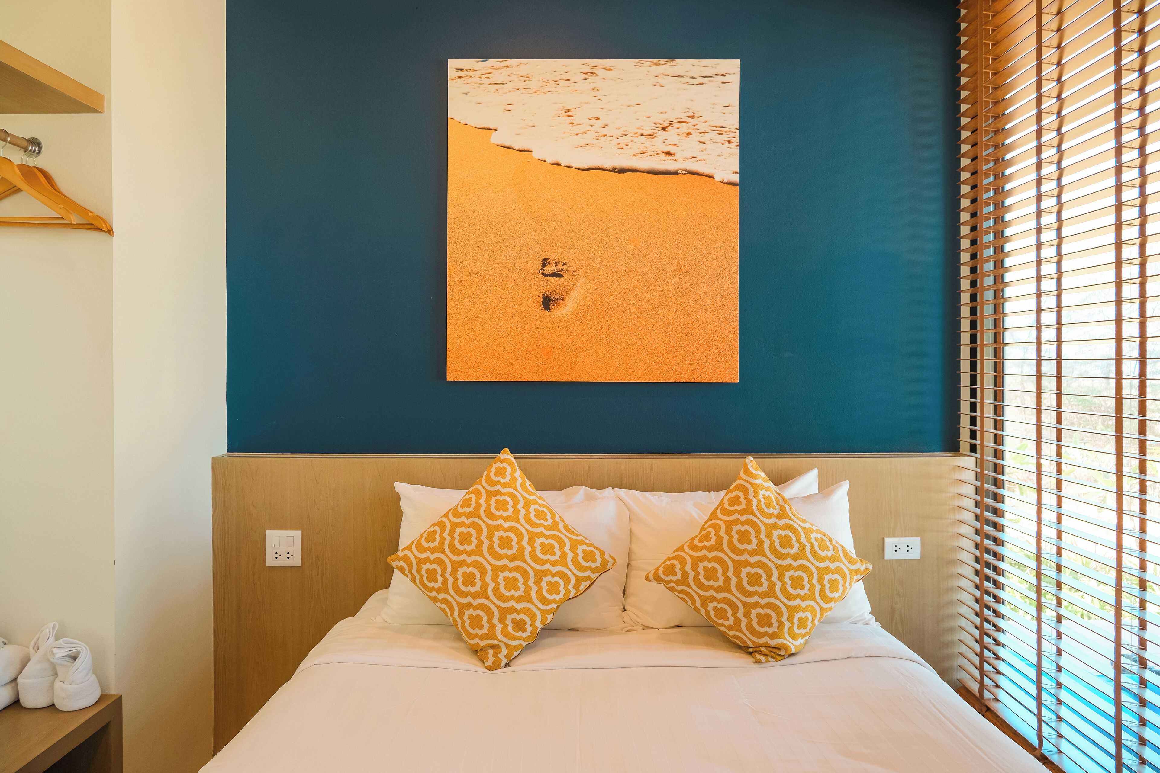 A hotel room with a bed, two patterned pillows, a wall-mounted artwork of a beach, and a window with blinds.