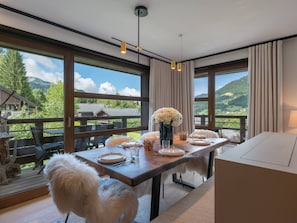 Uma área de refeições com uma mesa de madeira posta para dois, grandes janelas com vista para as montanhas e um cão branco e fofo sentado perto da mesa.