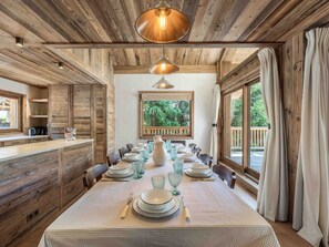 Interior de madeira com uma mesa de jantar posta para seis pessoas, uma janela ampla com vista para o verde e um lustre.