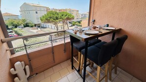 A balcony with a table set for two, chairs, a cactus decoration, and a view of buildings and greenery.
