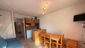 Un espacio habitable compacto con kitchenette, una zona de comedor con mesa y sillas de madera, un sofá y un televisor montado en la pared.