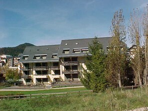 A modern building with a flat roof and multiple balconies, surrounded by greenery and a clear sky.