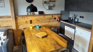 A compact kitchen with a wooden dining table, a black pendant light, and a wall-mounted shelf with decorative items.
