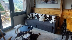 A cozy room with a grey sofa, a round table, and a large window offering a mountain view.