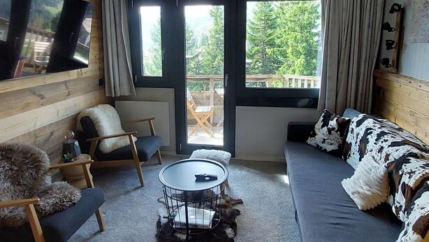 A cozy room with a wooden wall, a sofa, a chair, a small round table, and a TV mounted on the wall.