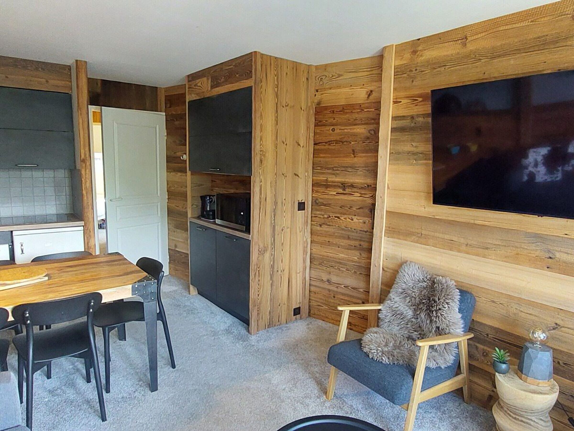 A compact living space with a wooden interior, a dining area with a table and chairs, a kitchenette, and a large flat-screen TV mounted on the wall.