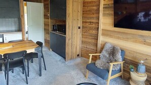 A compact living space with a wooden interior, a dining area with a table and chairs, a kitchenette, and a large flat-screen TV mounted on the wall.