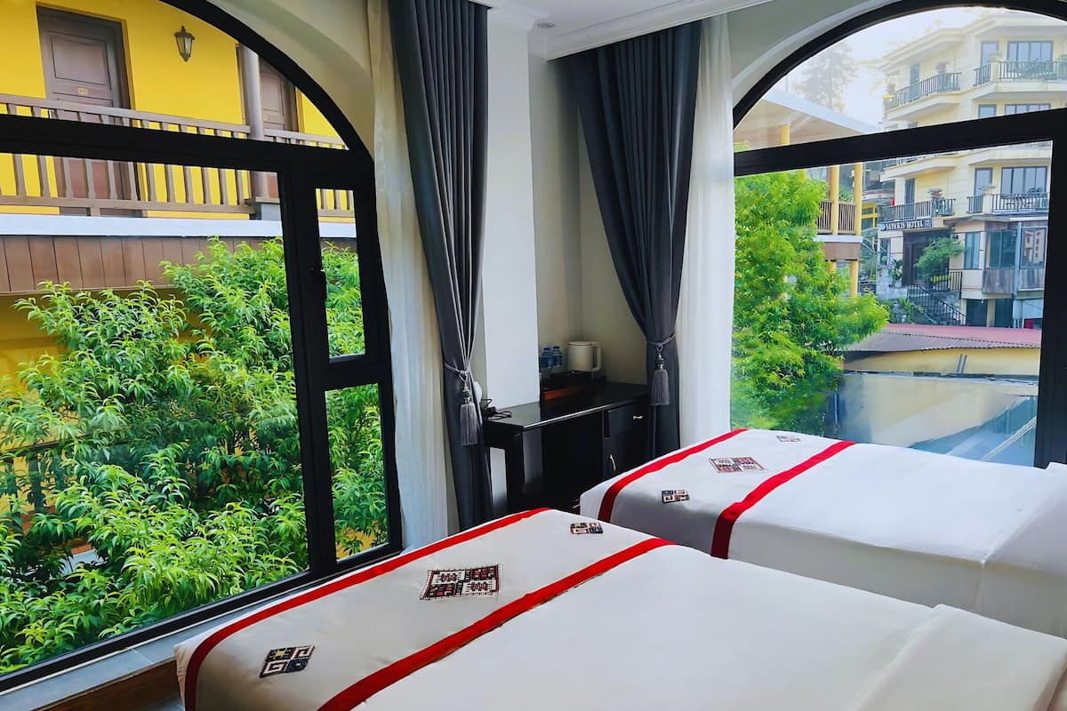 A hotel room with two beds, a small desk, and a window with a view of greenery and buildings.