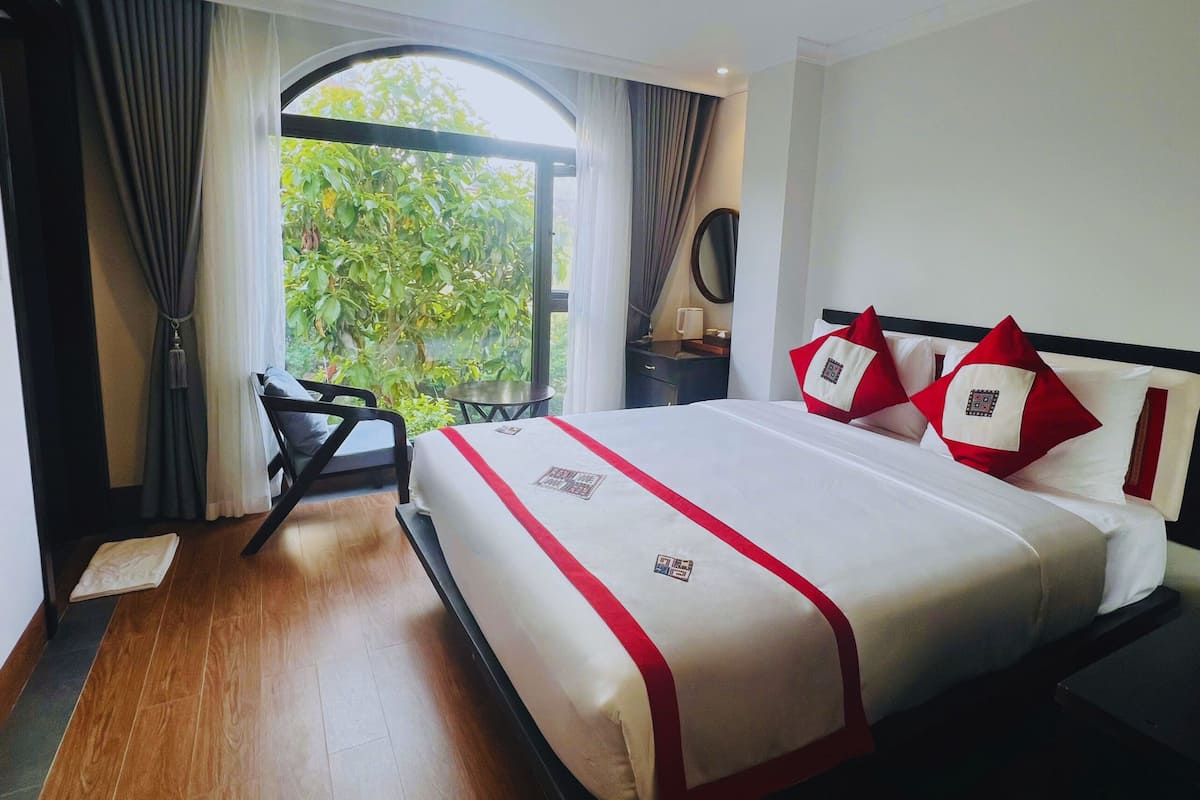 A hotel room with a large bed, a desk, and a view of greenery through a window.