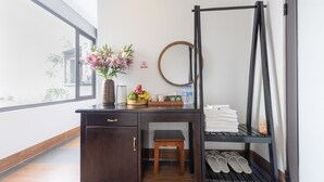 Une pièce avec un bureau en bois, un petit tabouret, une étagère à chaussures, un porte-rouleau de papier toilette suspendu et une fenêtre donnant sur des immeubles.