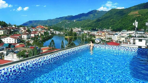Une piscine dont le bord est recouvert de carreaux, entourée de bâtiments et bordée par un lac, avec des montagnes en arrière-plan.