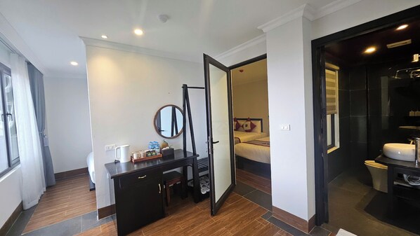 Ein Hotelzimmer mit Holzboden, einem Schreibtisch mit Spiegel, einem Bett mit Kissen sowie einem Badezimmer mit Waschbecken und Toilette.