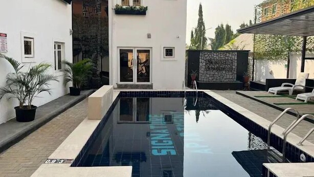 A swimming pool with a tiled deck, a white building with multiple windows, and a stone wall with greenery. - Quintessential Apartments By Signature (Abuja)