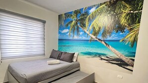 Ein Schlafzimmer mit Bett, einem Fenster mit Jalousien, Wandbild mit Strandmotiv und Holzboden.