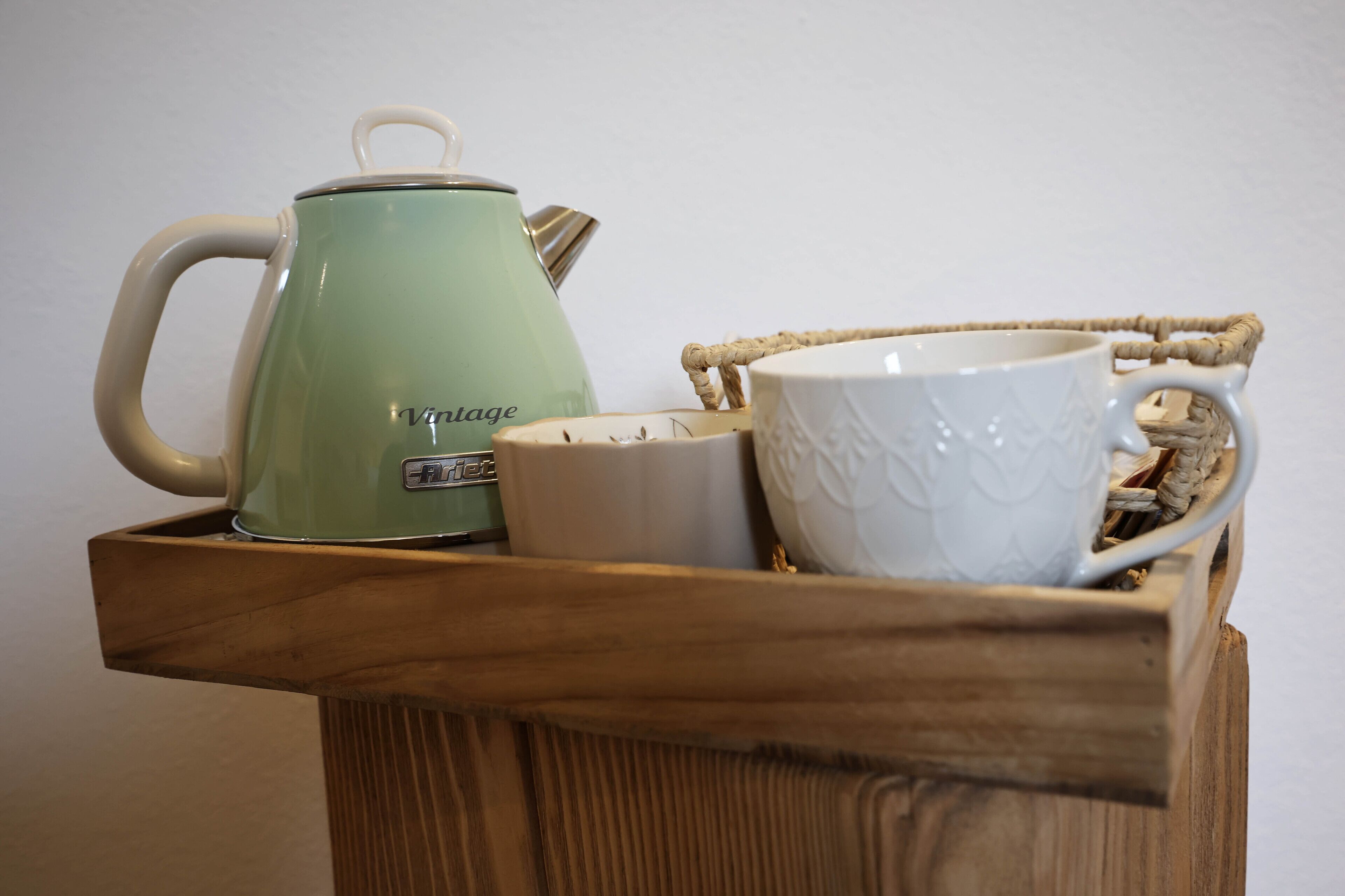 A wooden tray holding a green kettle, a white mug, and a white ceramic cup with a woven handle.