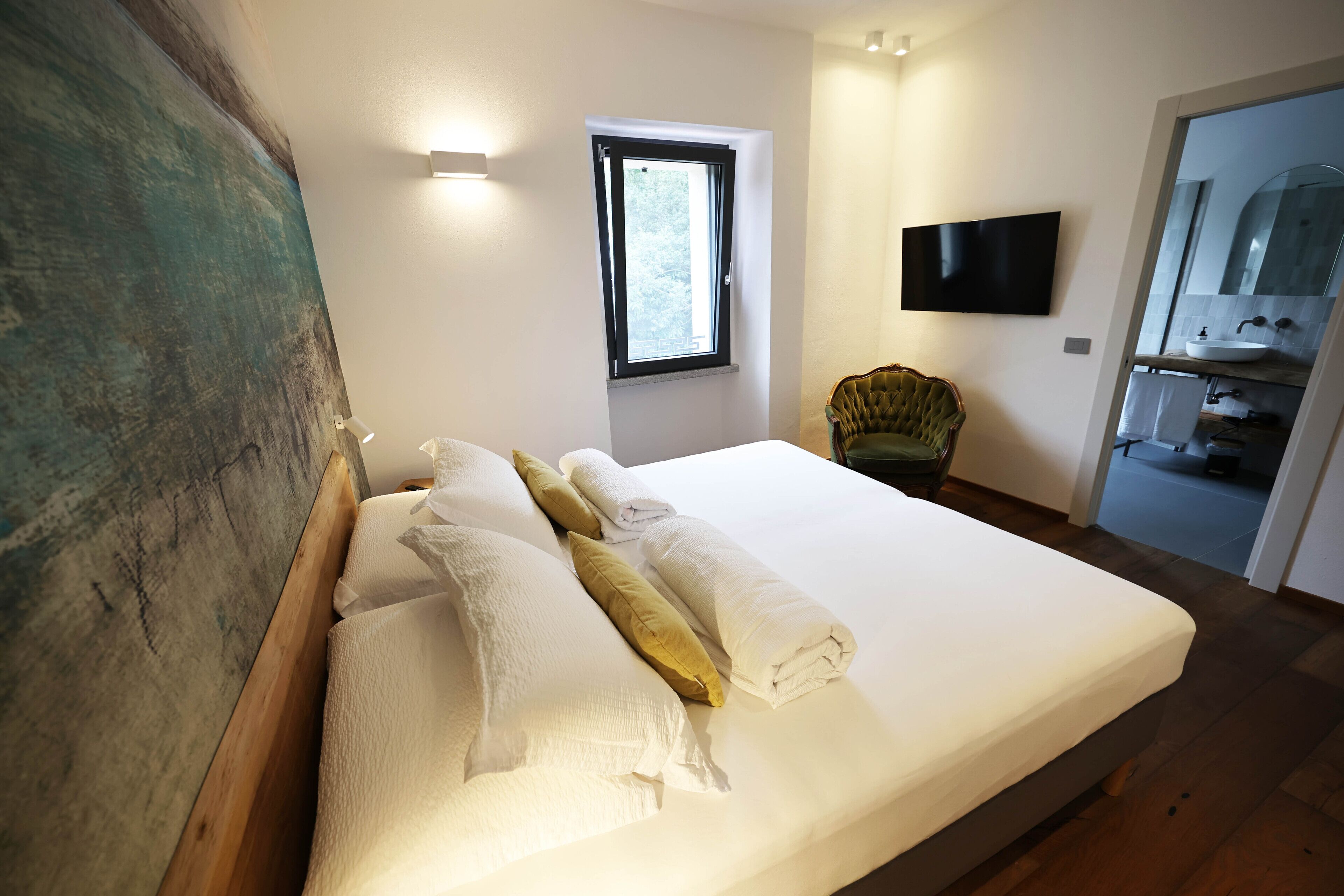 A neatly made bed with white and yellow pillows, a window with a view, a television on the wall, and a chair in the corner.
