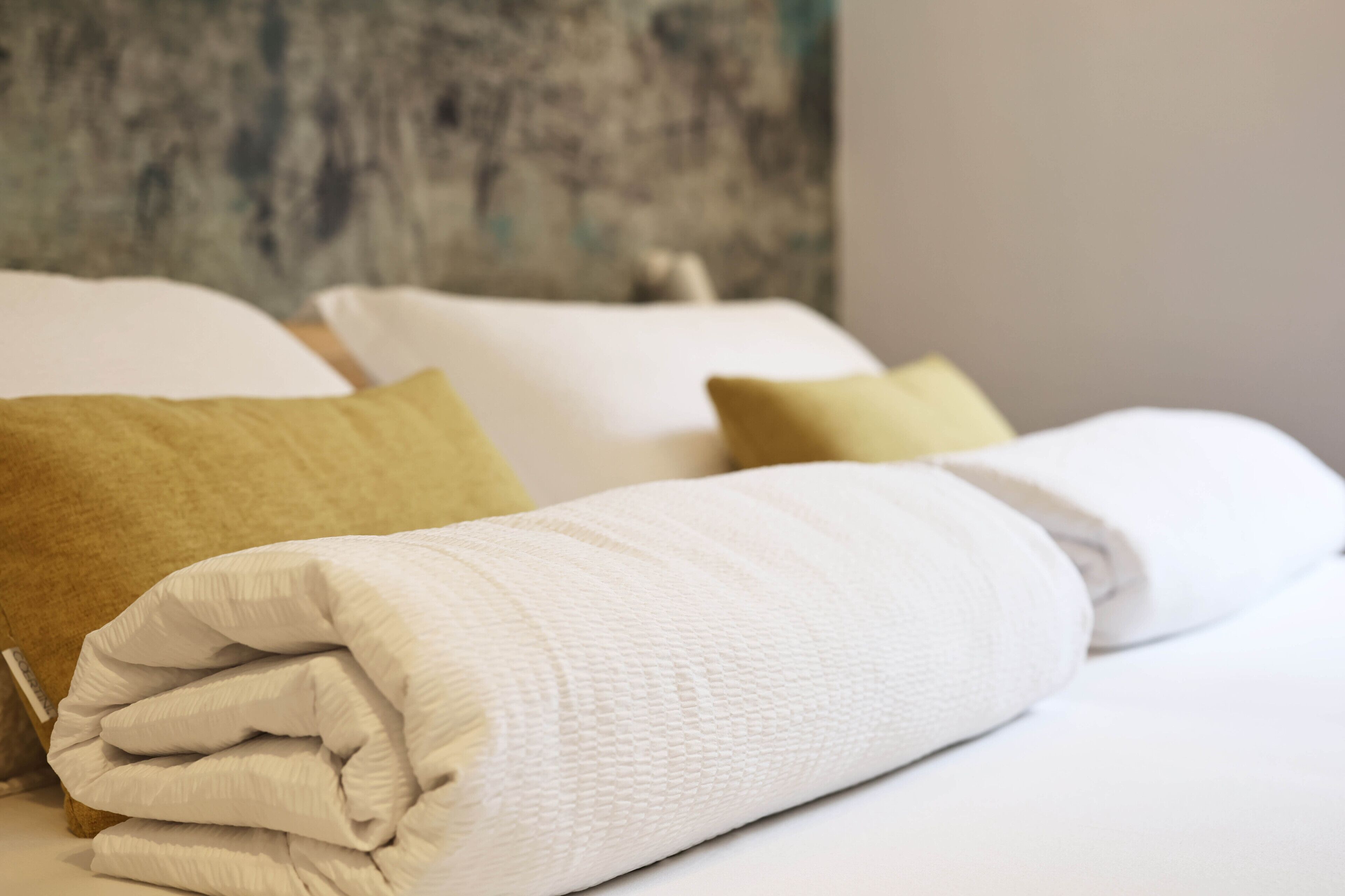 Una cama bien hecha con ropa de cama blanca y beige, incluyendo almohadas y un edredón enrollado.