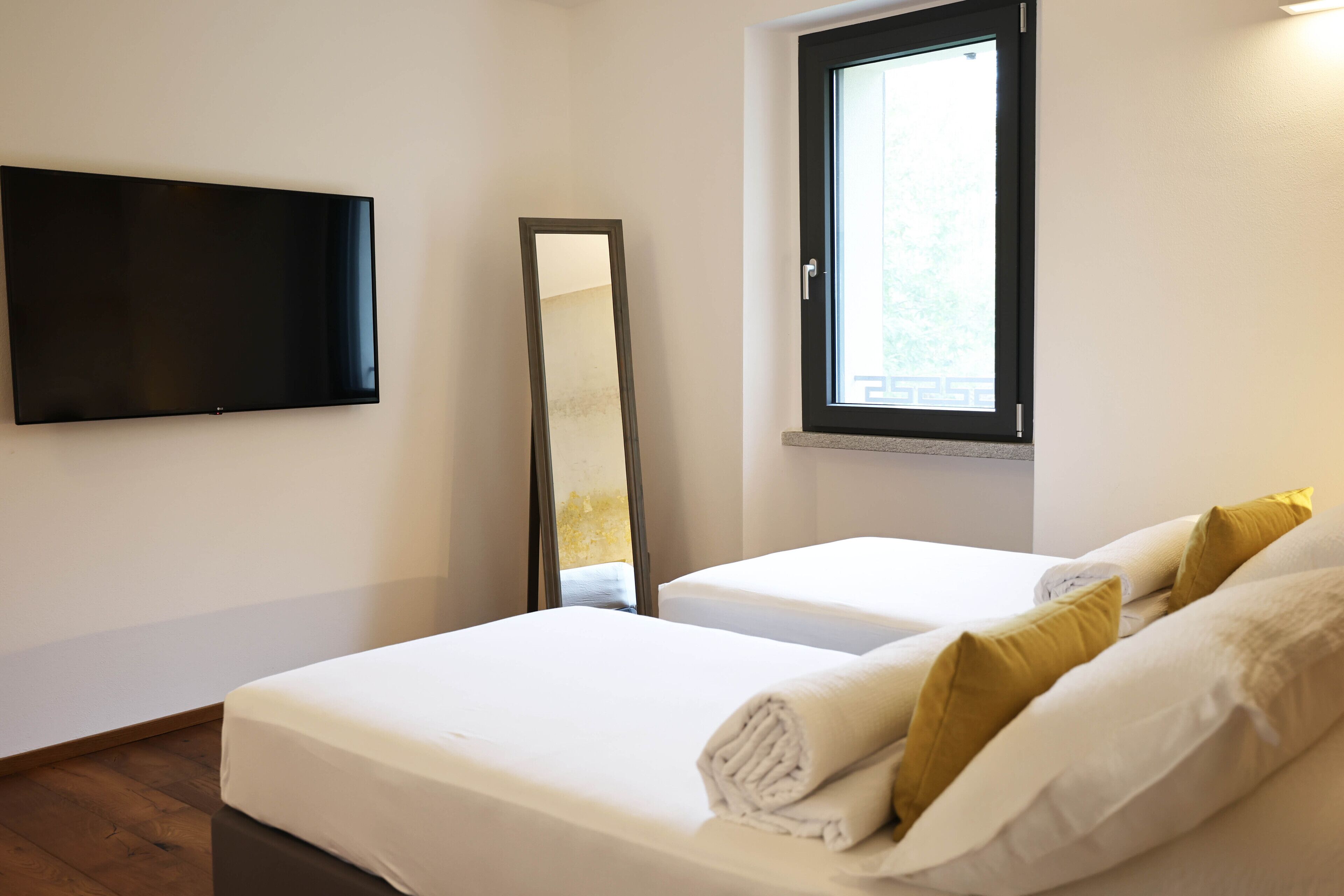 Quarto de hotel com televisão de tela plana na parede, um espelho grande, duas camas com roupa de cama branca e almofadas amarelas, e uma janela com vista para vegetação.