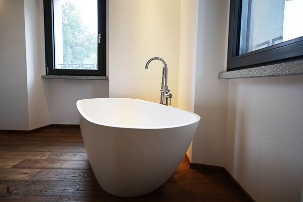 A modern bathroom with a freestanding bathtub, a window, and wooden flooring.