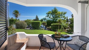 A balcony with a view of a well-maintained garden, a small round table with chairs, and a fan.
