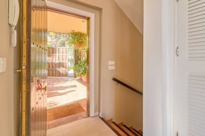 Ein Flur, der zu einer Tür mit Metallbeschlägen und Schlüsselloch führt, mit einem hölzernen Türrahmen, einer weißen Wand, einer Treppe mit Handlauf und Blick auf eine Terrasse mit Pflanzen und einem Holzzaun.