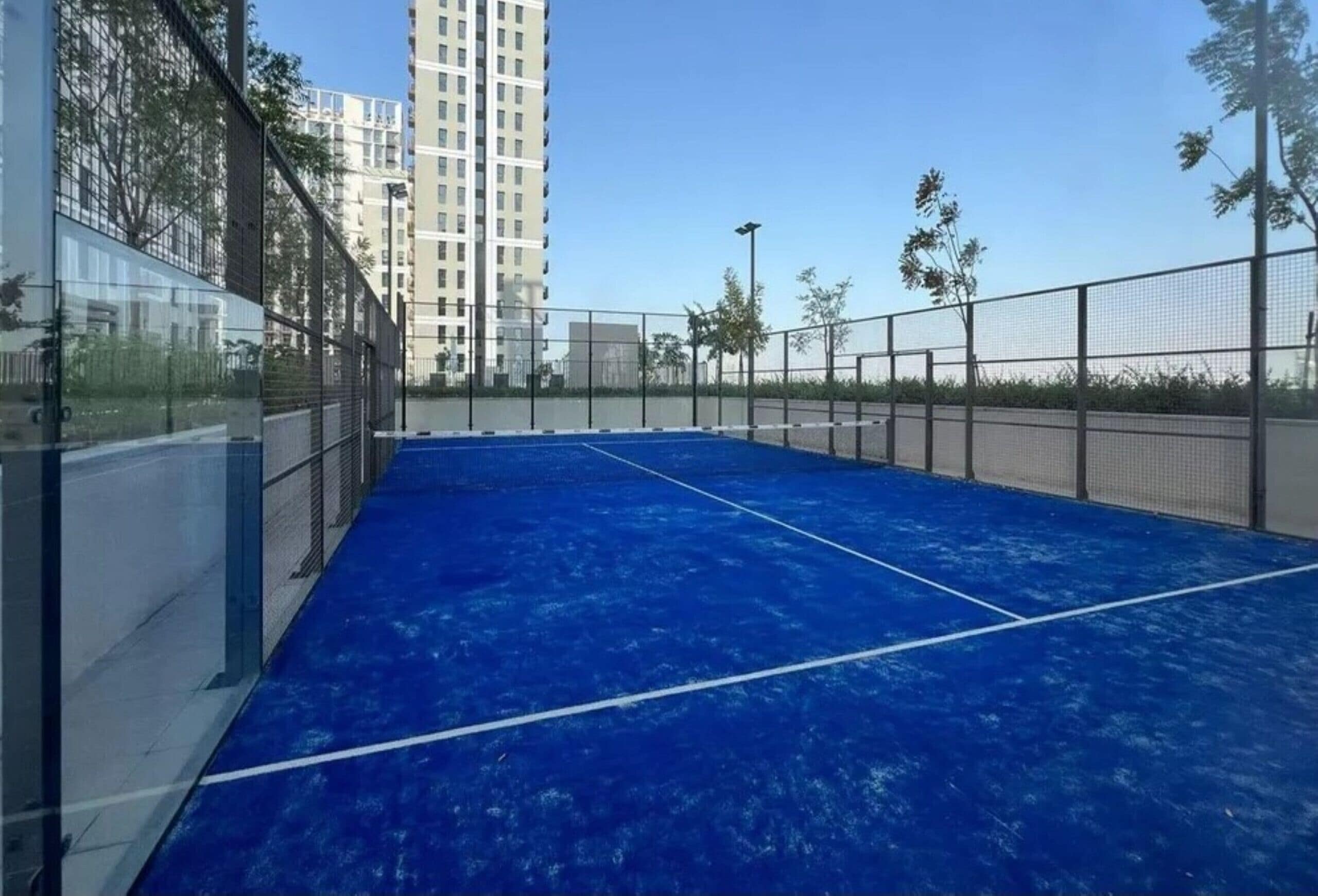 Una cancha de tenis con superficie azul, rodeada por una cerca y edificios altos al fondo.