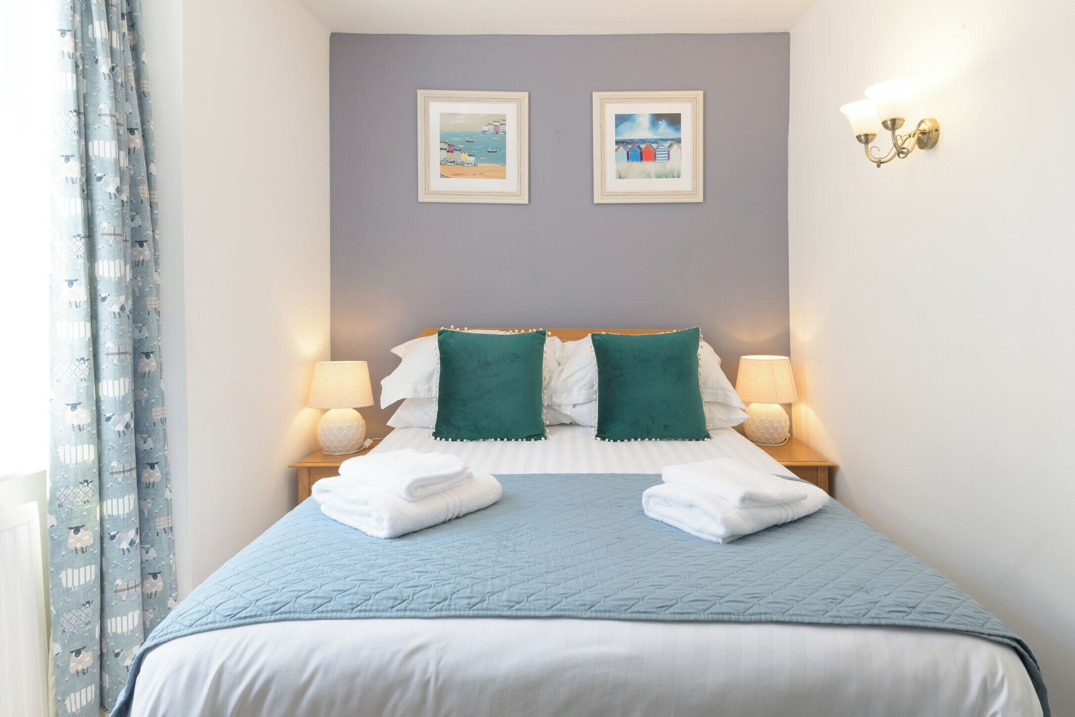 A neatly made bed with a blue quilt, two green pillows, and white linens. There are two framed pictures on the wall above the bed.