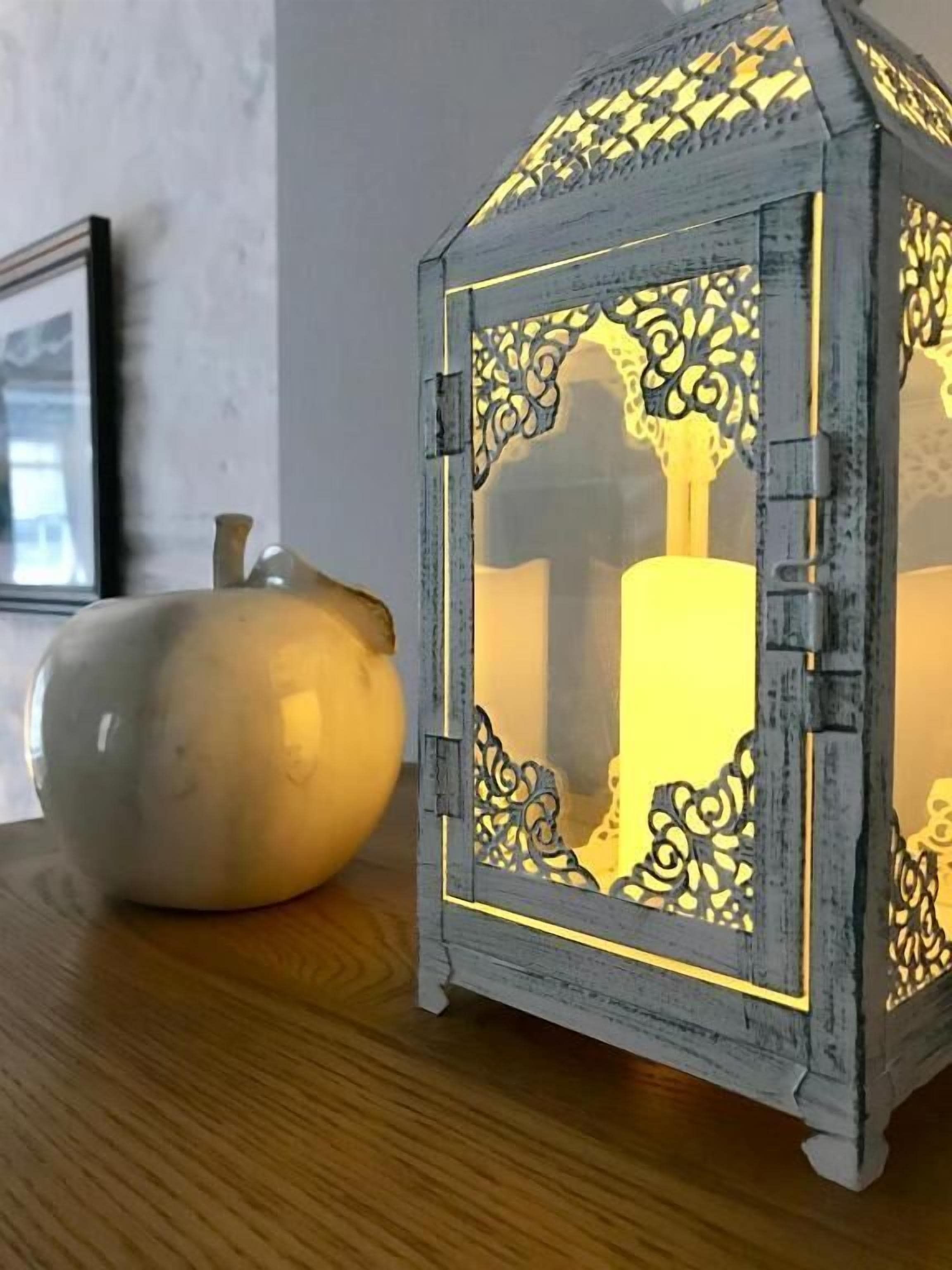 A small, ornate lantern with intricate cut-out designs and a candle inside, placed on a wooden surface next to a white ceramic object.